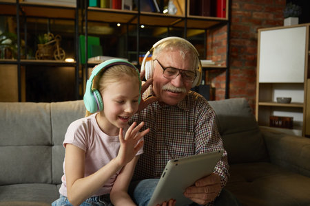 Smiling child girl and senior man, bot in headphones, sitting on couch and waving towards tablet, having online video call in cozy living room.の写真素材