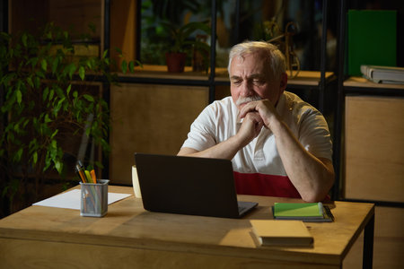 Focused elderly man sitting at wooden desk, using laptop and notebooks in quiet home office. Digital learning, modern technologiesの写真素材