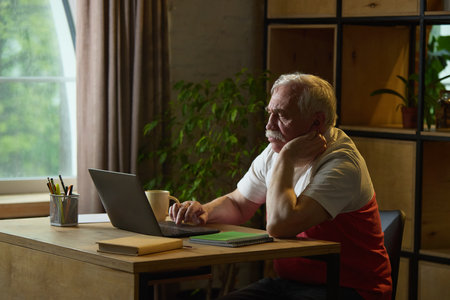 Focused elderly man with mustache sitting at wooden desk, looking intently at laptop with hand resting on chin, mug and notebooks nearbyの写真素材