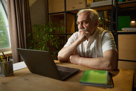 Elderly man with mustache sitting at wooden desk, looking thoughtfully at laptop with hand on chin, mug and notebooks nearby.の写真素材