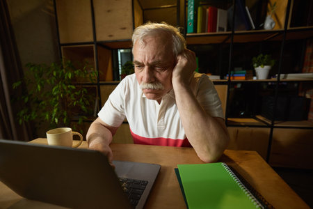 Elderly man with mustache sitting at wooden desk, looking down at laptop with one hand resting on his head, mug and notebooks nearby.の写真素材