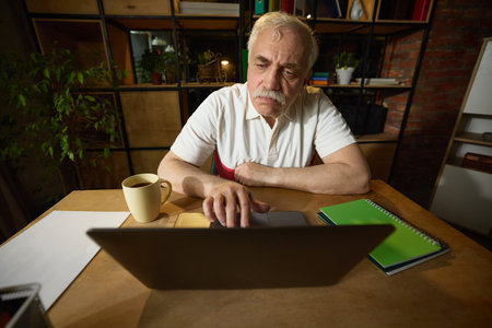 Elderly man with mustache sitting at wooden desk, looking at laptop with both hands on touchpad, mug and notebooks on the deskの写真素材