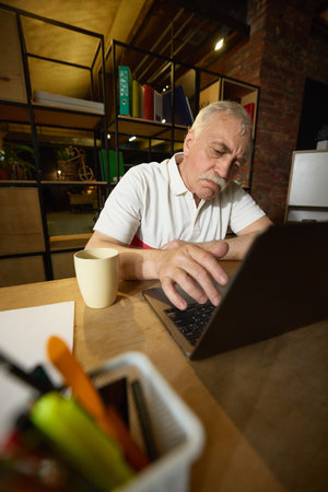 Elderly man with mustache sitting at wooden desk, looking down at laptop with hands on keyboard, mug on the desk.の写真素材