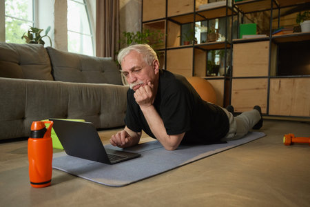 Elderly man lying on yoga mat and looking at laptop, with his chin resting on his hand. A water bottle and dumbbell on floor nearby.の写真素材