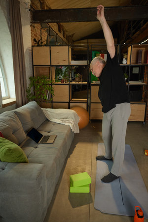 Elderly man standing on yoga mat with one arm raised overhead, performing exercise in living room. Laptop, yoga blocks visible.の写真素材