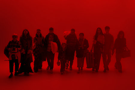 Crisis journey. Group of tired migrants walks through red neon glow with luggage and holding...