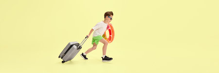 Boy in green shorts rushing forward with suitcase and colorful inflatable ring, expressing joy and freedom in motion against yellow background. Bannerの写真素材