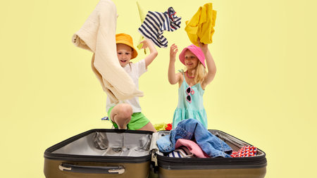 Boy and girl wearing bright hats and summer clothes throw colorful garments from suitcase into the air with joy, laughing together against yellow backdropの写真素材