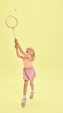 Girl in pink outfit jumps and strikes shuttlecock with badminton racket against yellow background, joyful expression and movement in motionの写真素材