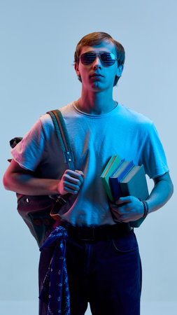 Young man with serious face holding colorful books and wearing shiny glasses standing in neon light against blue background. School vibeの写真素材