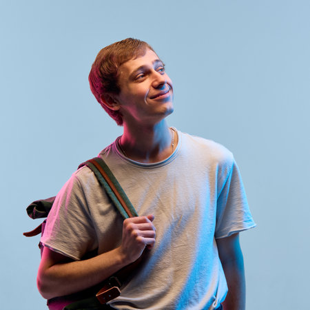 portrait of young man looking aside with soft dreamy smile in neon light against blue background as...