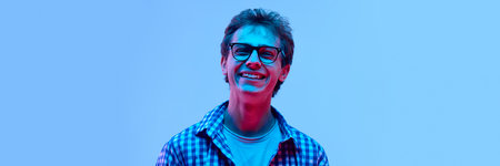 Happy young guy in glasses wearing checkered shirt laughing with closed eyes, looking cheerful and carefree in neon light against gradient backgroundの写真素材