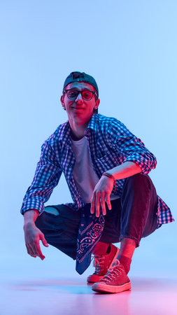 Stylish teenage boy in ripped jeans squatting down and smiling, wearing cap and glasses, showing relaxed vibe in neon light against gradient backgroundの写真素材