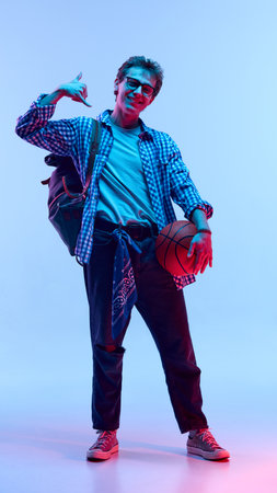Teenage boy in glasses and bandana holding basketball and showing phone hand gesture, standing confidently in stylish clothes in neon light against gradient background.の写真素材
