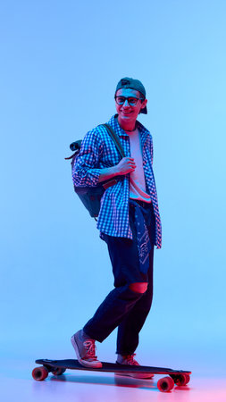 Trendy young man with glasses, cap and backpack riding longboard with smile, moving playfully and freely in neon light against gradient background.の写真素材
