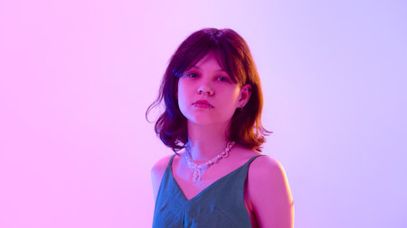 Serious teenage girl with piercing wearing green dress and pearl necklace posing under pink violet light against gradient backgroundの写真素材