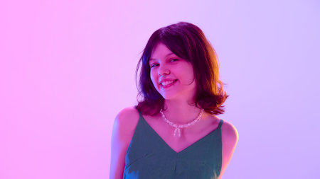 Playful girl in green dress smiling brightly, showing confidence under vibrant pink and violet light against gradient backgroundの写真素材
