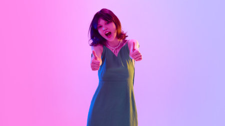 Excited teenage girl in green dress smiling and giving thumbs up under vibrant pink and violet neon light against gradient background.の写真素材