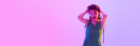 Shocked girl with open mouth holding head with hands under pink and violet neon lighting against...