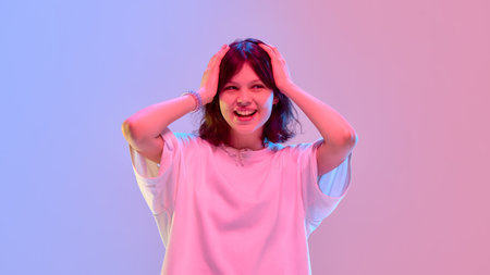 Surprised girl in white oversized shirt holding her head laughing under neon lights in emotional reaction against gradient backgroundの写真素材