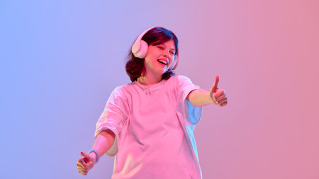 Energetic girl dancing with headphones showing thumbs up smiling confidently in glowing neon colors against gradient backgroundの写真素材