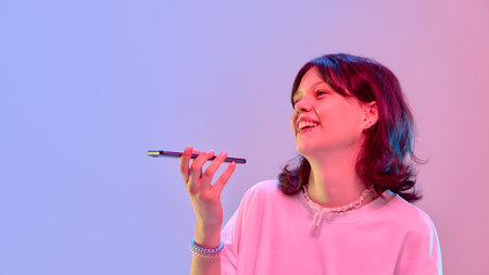 Happy girl laughing holding phone sideways during voice message under bright pink blue studio light against gradient background. Copy spaceの写真素材