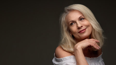 Senior woman smiling gently and resting chin on hand against dark background showing youthful glow and soft linesの写真素材