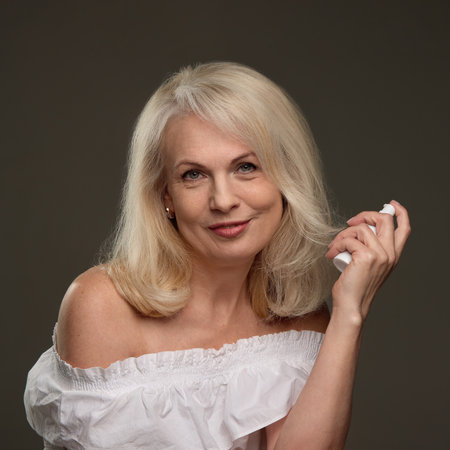 portrait of mature woman smiling softly holding spray bottle near face with gentle expression against dark backgroundの写真素材