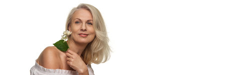 Mature woman holding flower near face with relaxed look showing radiant skin against white studio background with negative space. Bannerの写真素材