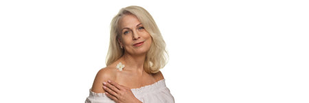 Mature woman touching shoulder with flower on skin smiling calmly against white studio background with negative space with negative space. Bannerの写真素材