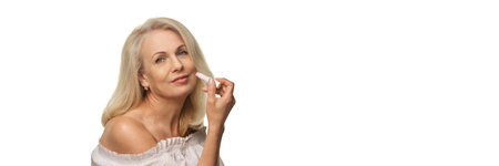 Elegant senior woman holding lip balm and smiling confidently with soft expression against white background in off-shoulder blouseの写真素材