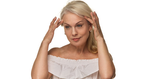 Senior woman gently touches temples while evaluating skin with focused expression against white background, showing beauty and self-acceptanceの写真素材