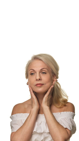 Graceful older woman presses hands to jawline, massaging face against white background, showing confident aging and daily facial careの写真素材