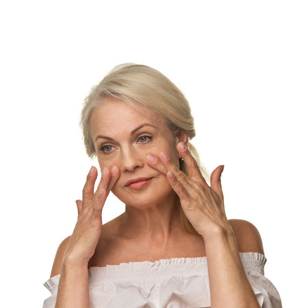 Elderly woman uses fingertips to massage skin under eyes against white background, reducing puffiness and supporting skincare routine with confidenceの写真素材