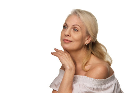 Graceful elderly woman looks upward with soft smile, gently holding hand under chin in thoughtful and radiant moment against white backgroundの写真素材