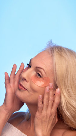 Senior woman gently adjusts peach-colored patches under her eyes, closing eyes and smiling softly while enjoying beauty routine.の写真素材