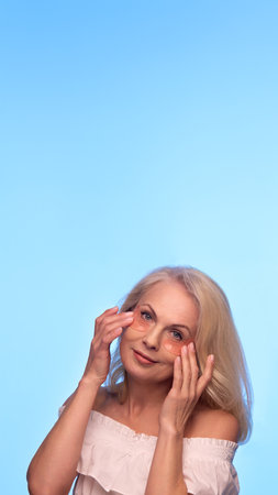 Mature woman gently adjusts peach-colored hydrogel patches under her eyes, closing eyes and smiling softly while enjoying beauty routineの写真素材