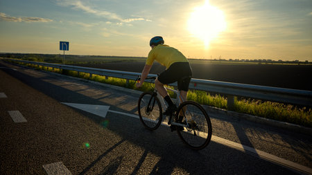 Rear view of cyclist riding toward horizon during golden hour, clear road and sun ahead with motion...