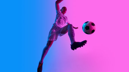 Female soccer player performs vertical jump with football in flight under striking pink-blue lights against gradient background. Low angle view.の写真素材