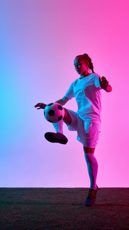 Woman football player balancing ball on raised leg with concentration and control under pink and blue light against gradient backdrop.の写真素材