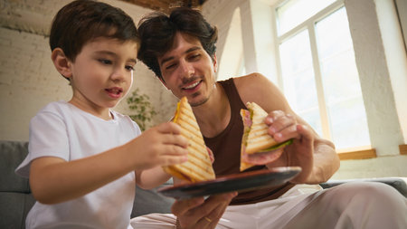 Father and young son enjoying grilled sandwiches together indoors on fathers dayの写真素材