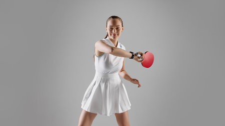 Smiling female table tennis player in white dress striking with red paddle on gray backgroundの写真素材