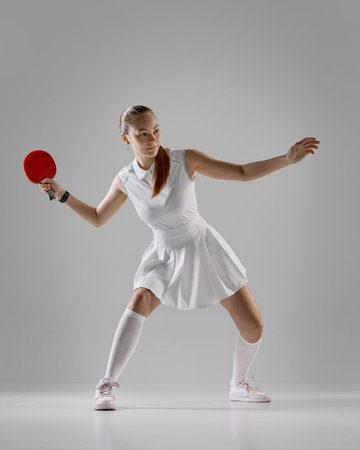 Focused female table tennis player in white outfit preparing to strike on gray backgroundの写真素材