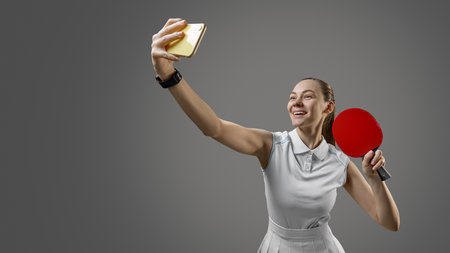 Cheerful female table tennis player posing for selfie with paddle on gray backgroundの写真素材