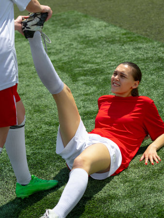 Female soccer player on ground stretching leg with teammate, intense expression in bright sunlight