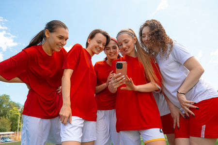 Group of young female soccer players in red and white jerseys smiling while looking at phone...