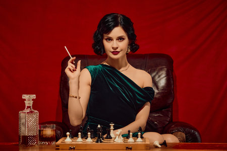 Woman in velvet dress holding cigarette and glass sitting at chessboard with intense expressionの写真素材