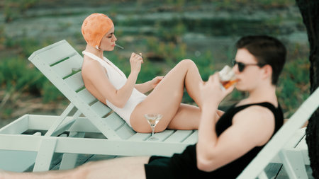Woman in white swimsuit by water with martini while man relaxes nearby