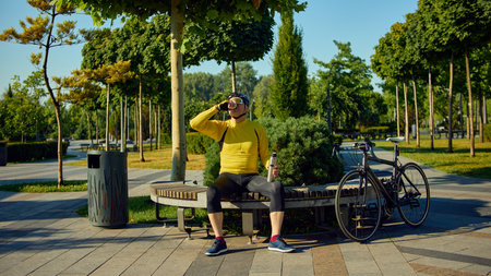 Elderly man in cycling gear drinking from thermos while sitting next to bikeの写真素材