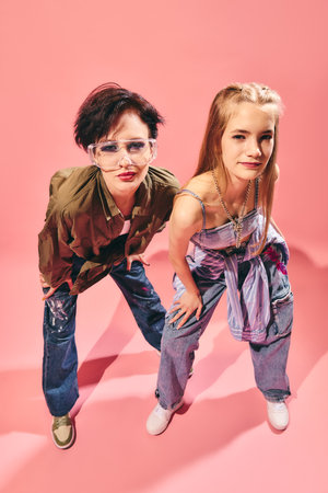 Two women in casual jeans and stylish tops posing together on pink background with confident...